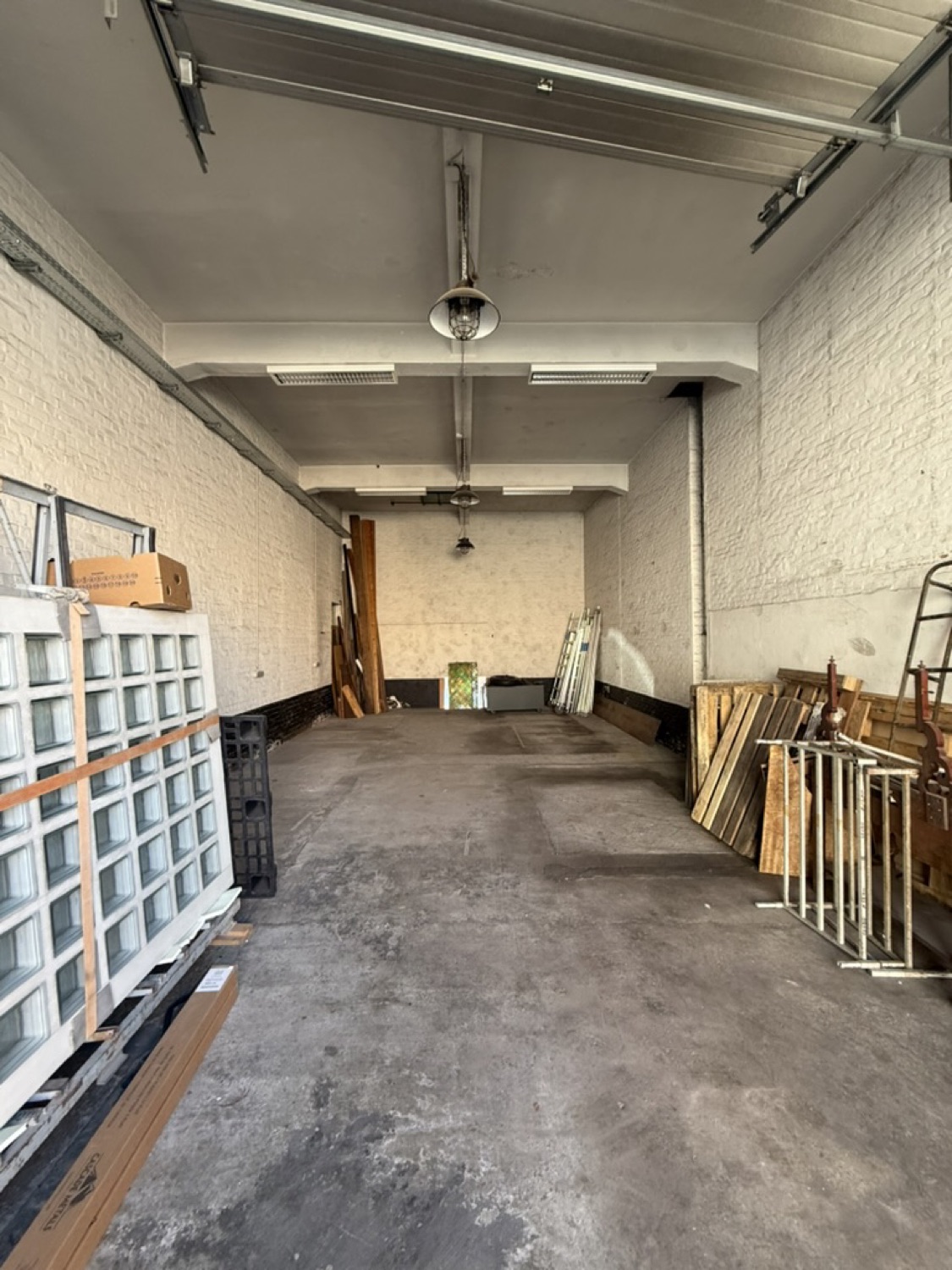 De garage van binnen: een lange, hoge ruimte met witte bakstenen muren, een hangende lamp en een betonnen vloer